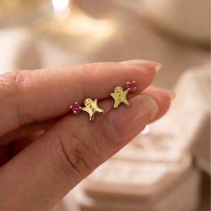 May include: A pair of gold stud earrings shaped like gingerbread figures, each featuring a small, round, vibrant pink gemstone set at the top corner. The gold figures have embossed facial details and a smooth, polished finish on the metal surface.