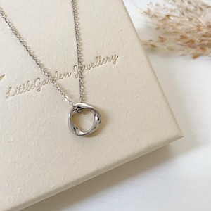 May include: A silver-toned necklace featuring a delicate chain and a small, irregular, circular pendant with a polished, reflective surface. The necklace is displayed on a cream-coloured box embossed with the text Little Garden Jewellery.