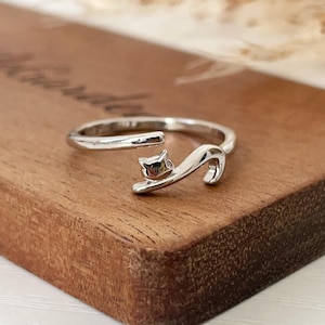May include: A silver-toned open ring featuring a stylised cat design. The band is smooth and polished, with one end shaped like a small cat head and the other end extending into a curved tail, creating an adjustable wrap-around appearance.