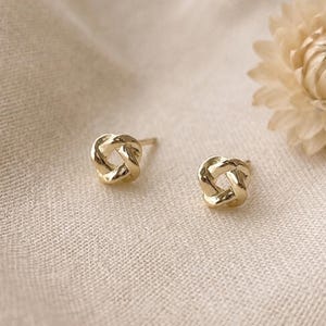 May include: A pair of gold stud earrings featuring a polished, intertwined knot design. Each earring has a small, rounded, four-loop structure that creates a continuous, knotted appearance. The metal has a bright, reflective finish.