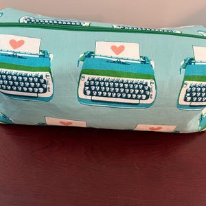 May include: A rectangular, light blue fabric bag with a green zipper. The bag is patterned with teal typewriters, each with a paper and a pink heart. The bag has a green zipper and green fabric handles on each end.