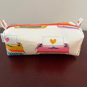 May include: A rectangular pencil case with a white background, featuring colorful typewriter illustrations. The typewriters are in shades of yellow, pink, and orange. The case has an orange zipper and small fabric loops on each end.