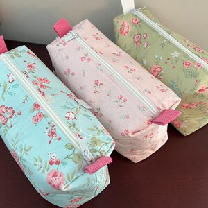 May include: Three rectangular fabric pouches with white zippers and pink fabric pull tabs. Each pouch features a floral pattern with small red and pink flowers on light blue, light pink, and light green backgrounds respectively.