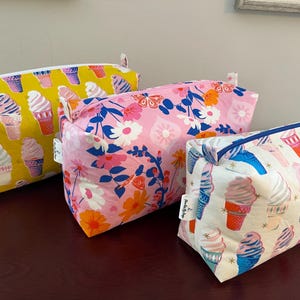 May include: Three rectangular fabric pouches with zipper closures. One is yellow with ice cream cones, another is pink with floral patterns, and the third is white with ice cream cones. All have a tag.