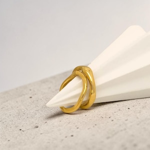 May include: A gold-colored ring with a double-band design. The ring is smooth and shiny, with a modern, minimalist aesthetic. The ring is resting on a white, folded paper structure, set against a neutral background.