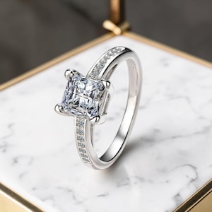 Può includere: Un anello d'argento con una grande pietra preziosa trasparente a taglio quadrato come elemento centrale. La fascia è inoltre ornata da pietre preziose chiare più piccole. L'anello è appoggiato su una superficie di marmo bianco e grigio.