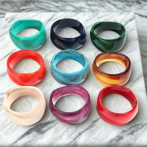 Peut inclure: Une collection de neuf bagues épaisses en résine disposées en une grille de trois par trois. Les bagues présentent des surfaces lisses et arrondies dans diverses couleurs unies et marbrées, notamment turquoise, bleu foncé, vert forêt, orange, rouge, beige et violet.
