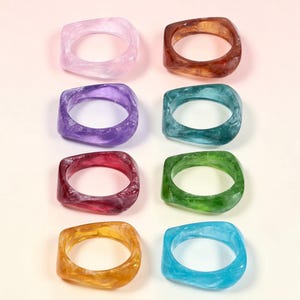 Può includere: Otto anelli quadrati in resina di vari colori, tra cui rosa, marrone, viola, verde acqua, rosso, verde, giallo e blu. Ogni anello ha un effetto marmorizzato, che aggiunge profondità al materiale traslucido. Gli anelli sono esposti su uno sfondo chiaro.