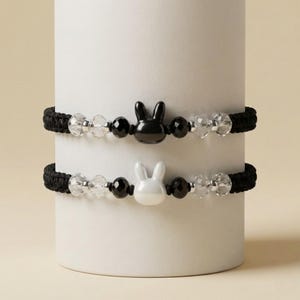 Puede incluir: Dos pulseras negras a juego con un dije de conejito. Cada pulsera presenta una banda negra tejida, cuentas transparentes y negras, y un dije de conejito. Un dije de conejito es negro, el otro es blanco.