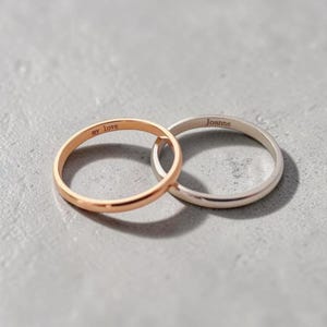 Könnte beinhalten: Zwei Eheringe, einer in Roségold und einer in Silber, liegen auf einer grauen Oberfläche. Der Roségoldring trägt die Inschrift "my love", der Silberring "Joanne".