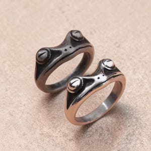 Puede incluir: Dos anillos plateados con forma de rana. Cada anillo presenta un diseño de rana con ojos grandes y redondos y una superficie texturizada. Los anillos se muestran sobre una superficie de tono neutro, mostrando su brillo metálico y forma única.