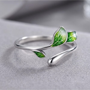 May include: A silver-toned open ring featuring a branch-like design with three small leaves. Two of the leaves are filled with a vibrant green enamel finish, while the third leaf is a plain, polished silver metal teardrop shape.