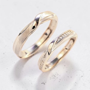 Könnte beinhalten: Zwei goldene Eheringe. Ein Ring hat eine strukturierte Oberfläche und ein verdrehtes Design. Der andere Ring hat ein ähnliches verdrehtes Design mit kleinen, klaren Steinen an einer Seite. Beide Ringe liegen auf einer weißen Oberfläche.