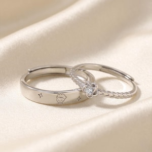 Könnte beinhalten: Zwei verstellbare Silberringe. Ein Ring zeigt eine kleine Cartoonfigur und Sterne. Der andere Ring hat ein Blumenmuster mit einem zentralen, klaren Stein und einem geflochtenen Band. Die Ringe liegen auf einem cremefarbenen Satinstoff.
