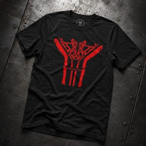 Op de afbeelding: Een antracietgrijs T-shirt met korte mouwen en een opvallende rode grafische print op de borst. Het ontwerp bestaat uit symmetrische, hoekige lijnen en geometrische vormen. Het label in de nek toont de letter M in een cirkelvormig logo.