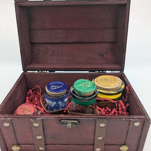 Może przedstawiać: Mała, otwarta drewniana skrzynia z ciemnoczerwonym wykończeniem, zawierająca trzy szklane słoiki z korkowymi zatyczkami. Słoiki mają niebieskie, zielone i żółte akcenty. Czerwone skrawki papieru wypełniają skrzynię.