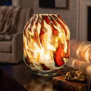 May include: A decorative glass vase with a unique pattern of red, orange, and white swirls. The vase is illuminated from within, creating a warm glow. It sits on a dark wooden surface, with a framed photo and a small brass figurine nearby.