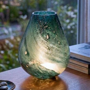 May include: A teardrop-shaped glass vase featuring a deep teal and green marbled pattern with subtle white flecks. The translucent glass has a smooth, polished finish and a wide, rounded base that tapers toward a narrow, open top opening.