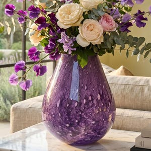 May include: A teardrop-shaped purple glass vase with a mottled white pattern, filled with a bouquet of cream-colored garden roses, vibrant purple sweet peas, and green eucalyptus stems, resting on a white marble surface next to stacked books.