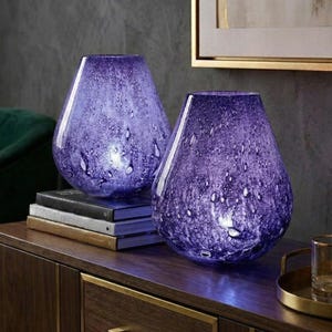 May include: Two purple glass vases with a speckled design. The vases are teardrop-shaped and sit on a dark wooden cabinet. The cabinet has gold-colored hardware. A green chair and artwork are in the background.