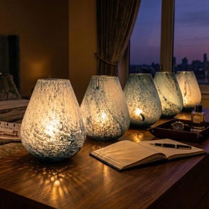 May include: A collection of four teardrop-shaped glass lamps with a speckled blue and white pattern. The lamps are illuminated from within, casting a warm glow on a wooden surface. Each lamp features a unique mottled texture in shades of blue.