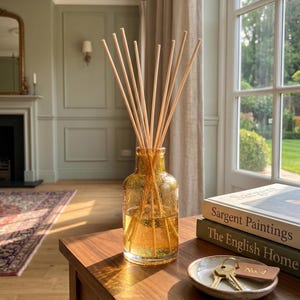 May include: A glass reed diffuser bottle in a warm amber hue with a textured, bubbly surface, containing a clear liquid and several light-coloured wooden reeds. Beside it sits a small ceramic dish holding a set of metal keys with a leather tag.
