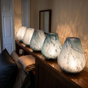 May include: Five teardrop-shaped glass lamps arranged in a row on a wooden surface. Each lamp features a unique mottled texture with varying shades of white, gray, and blue, emitting a soft, warm internal glow that illuminates the surrounding area.