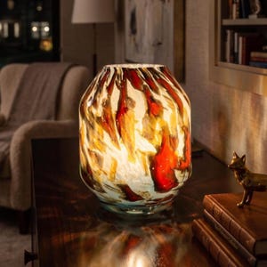 May include: A glass table lamp with a rounded, barrel-shaped body featuring a swirling, marbled pattern of deep red, amber, and cream colors. The lamp emits a warm, diffused glow from within, highlighting the translucent glass texture.