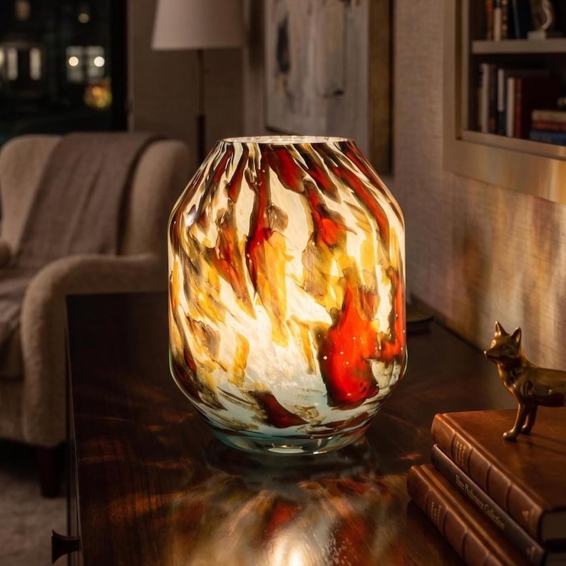 May include: A glass table lamp with a rounded, barrel-shaped body featuring a swirling, marbled pattern of deep red, amber, and cream colors. The lamp emits a warm, diffused glow from within, highlighting the translucent glass texture.