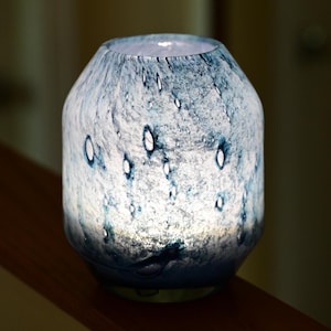 Puede incluir: Una l&aacute;mpara cil&iacute;ndrica de vidrio con un acabado moteado en azul y blanco, que presenta una superficie texturizada con hendiduras claras en forma de gotas de lluvia. La l&aacute;mpara emite un suave resplandor interno que resalta los patrones.