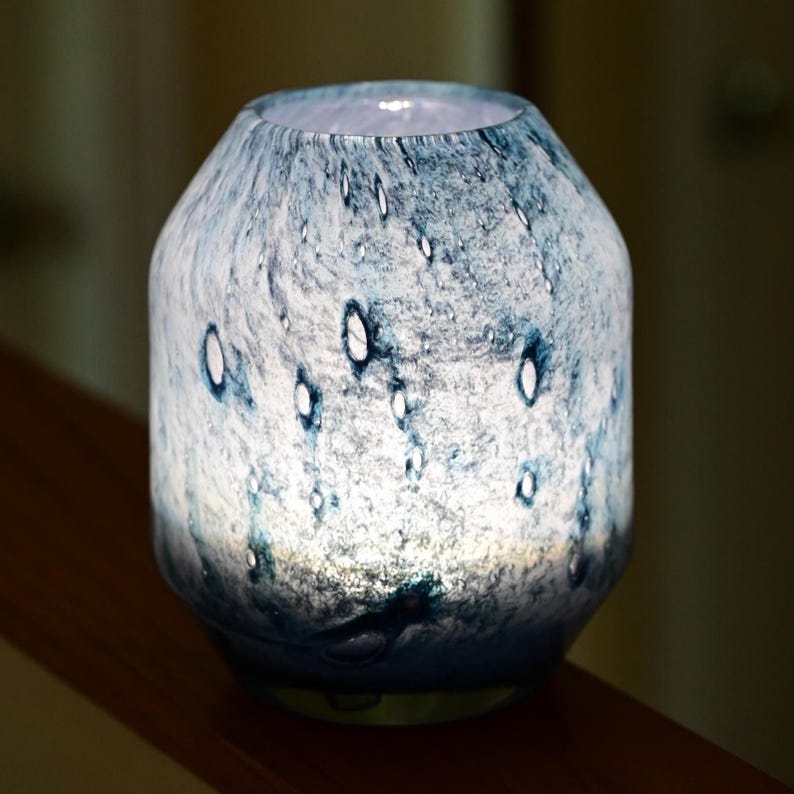 Puede incluir: Una l&aacute;mpara cil&iacute;ndrica de vidrio con un acabado moteado en azul y blanco, que presenta una superficie texturizada con hendiduras claras en forma de gotas de lluvia. La l&aacute;mpara emite un suave resplandor interno que resalta los patrones.