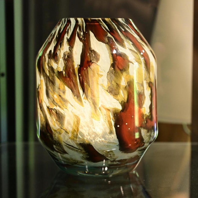 May include: A glass vase with a wide, rounded body and a narrow opening. The surface features a swirling, abstract pattern of deep red, amber, and dark brown streaks layered over a translucent, off-white base, creating a marbled effect.