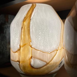 May include: A glass vase featuring a rounded, bulbous shape with a textured, opaque white surface. The design is accented by swirling, translucent amber-colored glass ribbons that create organic, flowing patterns across the entire exterior.