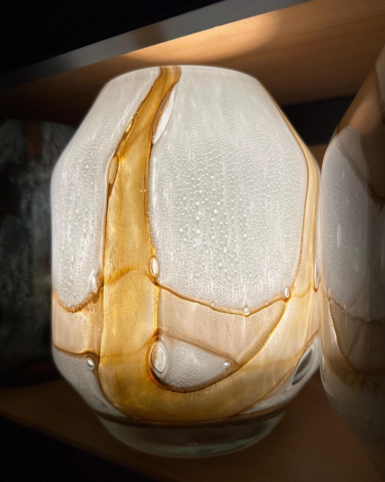 May include: A glass vase featuring a rounded, bulbous shape with a textured, opaque white surface. The design is accented by swirling, translucent amber-colored glass ribbons that create organic, flowing patterns across the entire exterior.