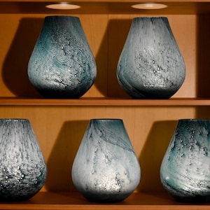 May include: Five teardrop-shaped glass vases arranged on two wooden shelves. Each vase features a mottled, swirling pattern in shades of teal, blue, and white, with a textured surface containing small air bubbles throughout the glass material.