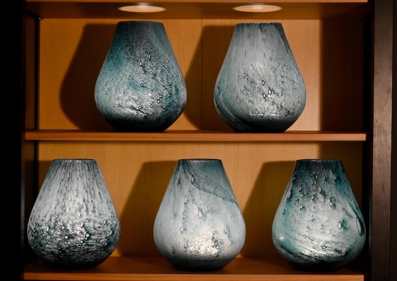 May include: Five teardrop-shaped glass vases arranged on two wooden shelves. Each vase features a mottled, swirling pattern in shades of teal, blue, and white, with a textured surface containing small air bubbles throughout the glass material.