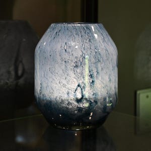 Puede incluir: Un florero de vidrio con un patr&oacute;n moteado en blanco y varios tonos de azul. El recipiente tiene un cuerpo ancho y redondeado que se estrecha hacia una abertura m&aacute;s peque&ntilde;a en la parte superior, con un acabado liso y un degradado de azul oscuro.