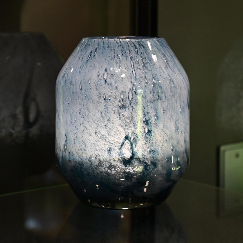 Puede incluir: Un florero de vidrio con un patr&oacute;n moteado en blanco y varios tonos de azul. El recipiente tiene un cuerpo ancho y redondeado que se estrecha hacia una abertura m&aacute;s peque&ntilde;a en la parte superior, con un acabado liso y un degradado de azul oscuro.