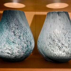 May include: Two teardrop-shaped glass vases featuring a mottled blue and white finish with a textured, bubbly surface. The vases have a wide base that tapers toward a narrow opening, displaying a blend of deep teal and pale icy blue tones.