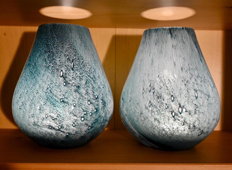 May include: Two teardrop-shaped glass vases featuring a mottled blue and white finish with a textured, bubbly surface. The vases have a wide base that tapers toward a narrow opening, displaying a blend of deep teal and pale icy blue tones.