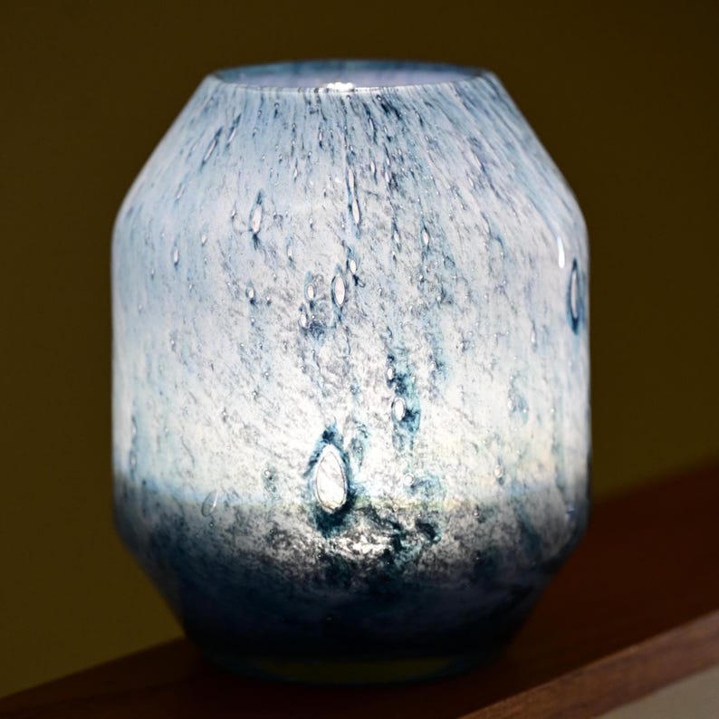 Puede incluir: Un florero cil&iacute;ndrico de vidrio con un patr&oacute;n moteado en azul y blanco con efecto degradado, que transita desde una parte superior clara y transl&uacute;cida hacia una base azul marino opaca y profunda, con peque&ntilde;as burbujas de aire.