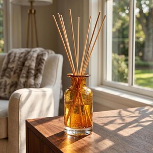 Op de afbeelding: Een doorzichtige amberkleurige glazen geurstokjesfles met een korte hals en een ronde basis, gevuld met een goudkleurige vloeistof. Verschillende dunne, lichtgekleurde houten stokjes zijn in de fles gestoken en waaieren uit vanuit de opening.