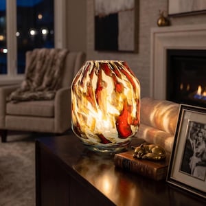 May include: A cylindrical glass lamp with a swirling pattern of deep red, amber, and white colors. The lamp emits a warm, glowing light from within, illuminating the intricate, marbled glass texture across its rounded, wide-mouthed body.