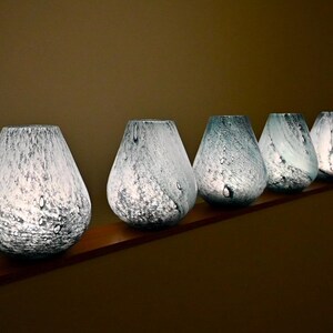 May include: A row of five teardrop-shaped glass vases featuring a mottled white and light blue speckled pattern. Each vase has a wide base that tapers toward a circular opening at the top, with a translucent quality that allows light to pass through.