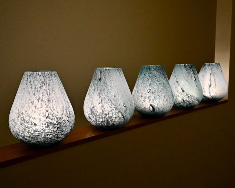 May include: A row of five teardrop-shaped glass vases featuring a mottled white and light blue speckled pattern. Each vase has a wide base that tapers toward a circular opening at the top, with a translucent quality that allows light to pass through.