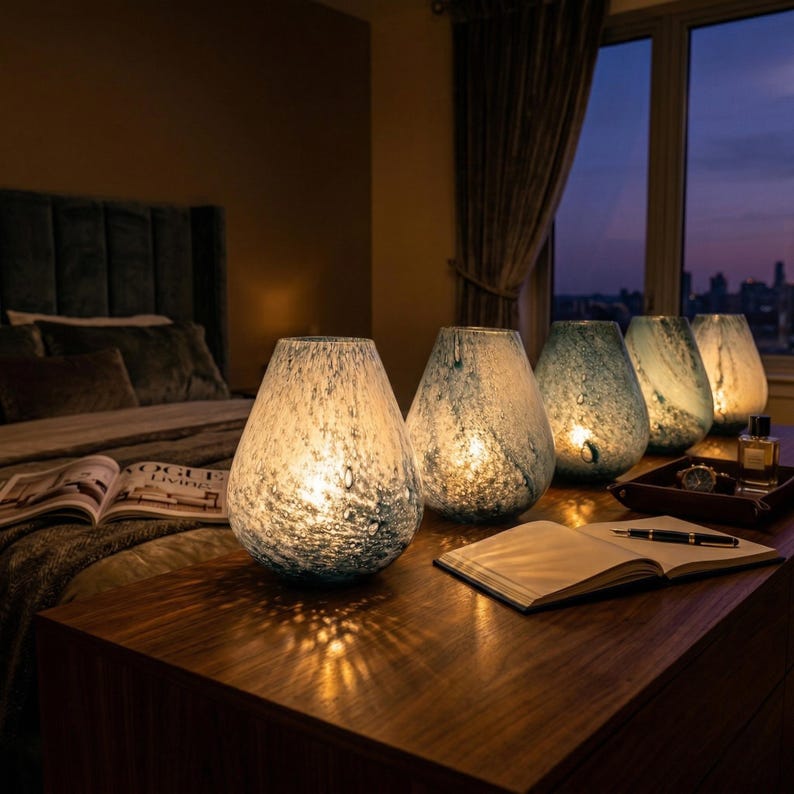 May include: A collection of four teardrop-shaped glass lamps with a speckled blue and white pattern. The lamps are illuminated from within, casting a warm glow on a wooden surface. Each lamp features a unique mottled texture in shades of blue.