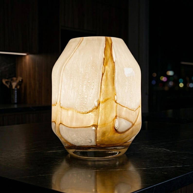 May include: A glass vase featuring a rounded, faceted silhouette with a swirling pattern of translucent cream and amber-colored glass. The surface displays a textured, bubbled appearance throughout, with distinct golden-brown veins running vertically.