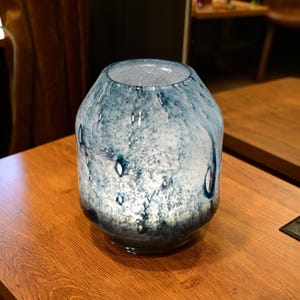 Puede incluir: Un florero de vidrio redondeado con una mezcla jaspeada de colores azul profundo y blanco. La superficie tiene una apariencia texturizada con burbujas de aire visibles incrustadas en el vidrio, creando un efecto transl&uacute;cido en el cuerpo.