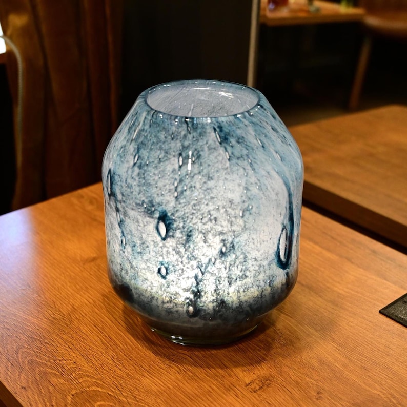 Puede incluir: Un florero de vidrio redondeado con una mezcla jaspeada de colores azul profundo y blanco. La superficie tiene una apariencia texturizada con burbujas de aire visibles incrustadas en el vidrio, creando un efecto transl&uacute;cido en el cuerpo.
