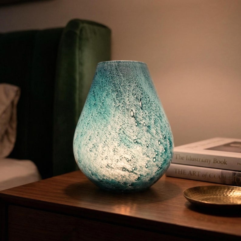 May include: A teardrop-shaped glass table lamp featuring a mottled teal and white finish with embedded air bubbles. The lamp emits a soft, warm glow from within, showcasing a textured surface that transitions from deep blue-green to white.